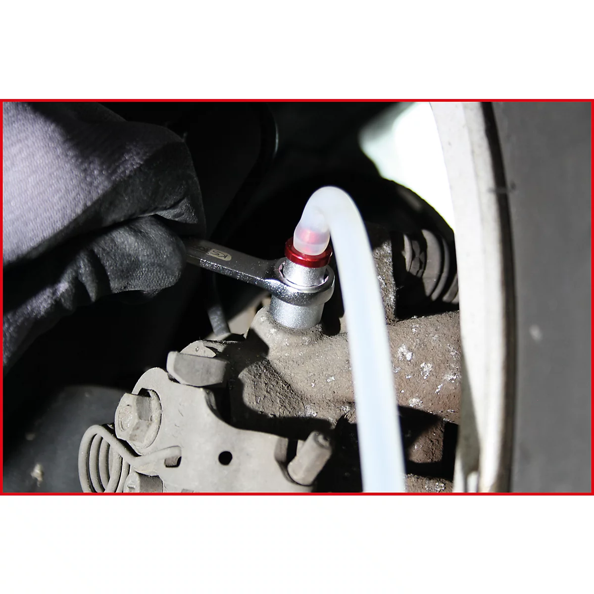 Ein Mann verschraubt in Nahaufnahme eine Bremsleitung mit einem Schraubenschlüssel, an dem ein transparenter Schlauch befestigt ist.